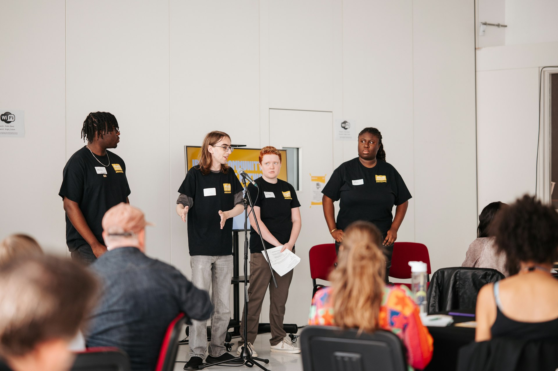 Three young people in black "The Power of Youth" t-shirts stand at a microphone presenting to a seated audience in a white-walled room